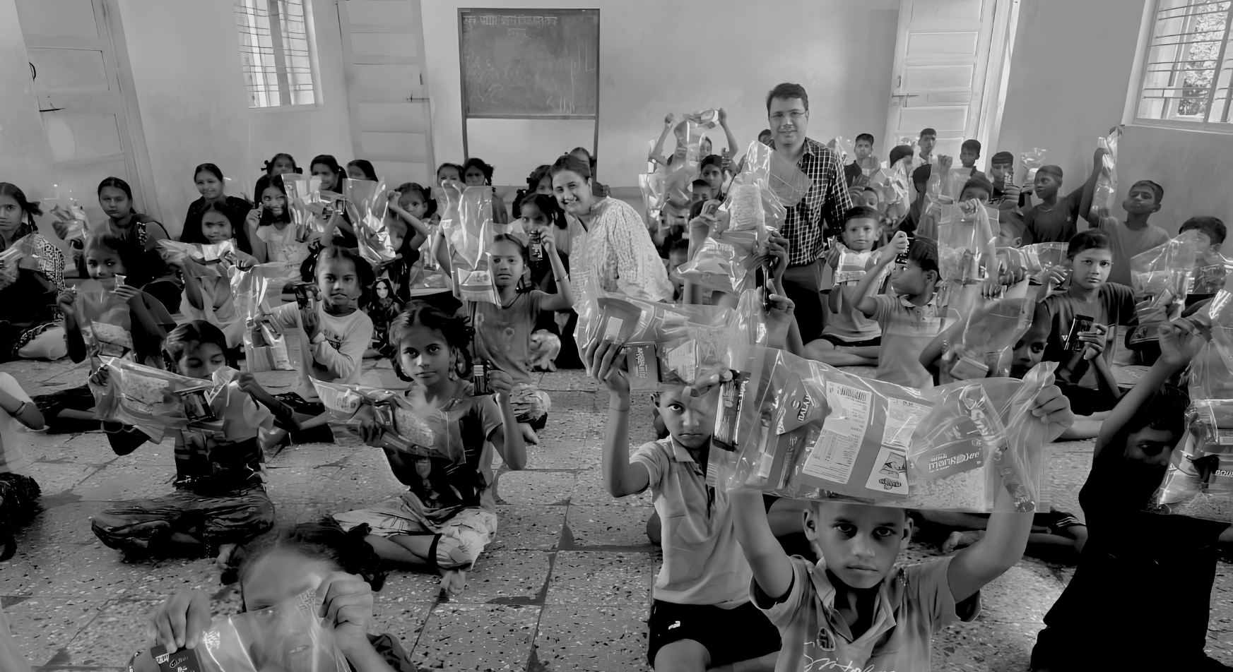 Children seated on a classroom floor holding up gift packets received during an outreach visit